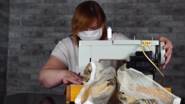 Dikiş makinesi kullanan yetişkin kadın perdesini kapat. Tıbbi maskeli genç terzi evde dikiş makinesi üzerinde çalışıyor..