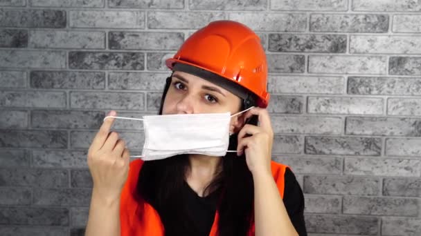 Trabalhadora da construção feminina em macacões colocando máscara médica no rosto no fundo da parede de tijolo cinza — Vídeo de Stock