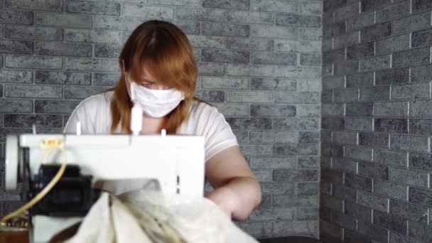 Nahaufnahme einer erwachsenen Frau, die mit einer Nähmaschine Gardinen näht. Junge Näherin in medizinischer Maske arbeitet zu Hause an Nähmaschine. — Stockvideo