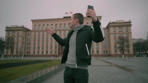 Een jonge knappe man in casual kleding dirigeert een live uitzending in zijn mobiele telefoon op een plein in het centrum van de stad. — Stockvideo