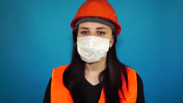 Vrouwelijke bouwvakker in overall verwijdert medisch masker van gezicht op blauwe achtergrond. — Stockvideo