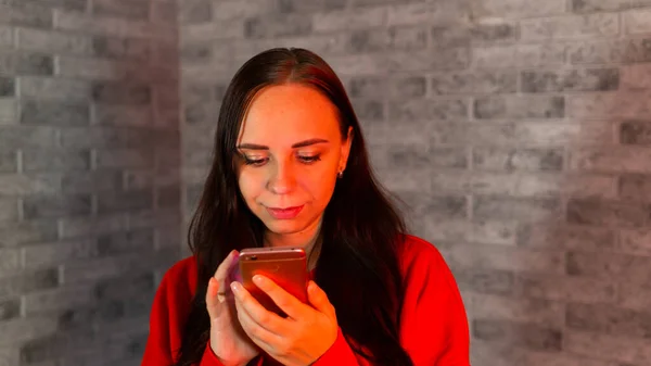 Mulher bonita com capuz vermelho lançando notícias de coronavírus no telefone celular, de pé sobre fundo de tijolo cinza . — Fotografia de Stock