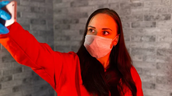 Молодая женщина в медицинской маске и перчатках делает селфи на смартфоне, стоя на сером кирпичном фоне. Портрет женщины, проводящей выходные дома и фотографирующей себя на мобильный телефон . — стоковое фото
