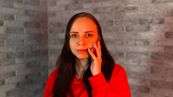 Csinos fiatal nő piros kapucnis beszél koronavírus a mobiltelefon, álló szürke tégla háttér. — Stock Fotó