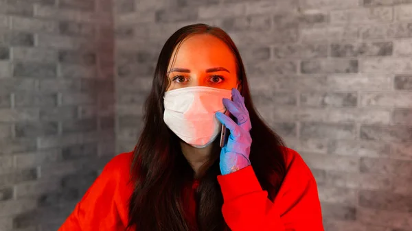 Красивая молодая женщина в медицинской маске и перчатках говорит о коронавирусе на мобильном телефоне, стоя на сером кирпичном фоне . — стоковое фото