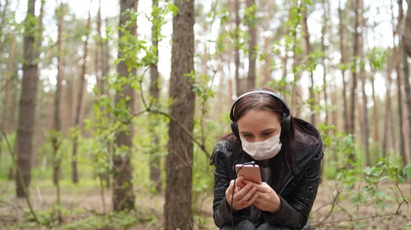 Nahaufnahme einer jungen Frau in medizinischer Maske, die Musik hört und im Handy Nachrichten durchblättert, auf einem Baumstamm im Wald sitzt. — Stockfoto