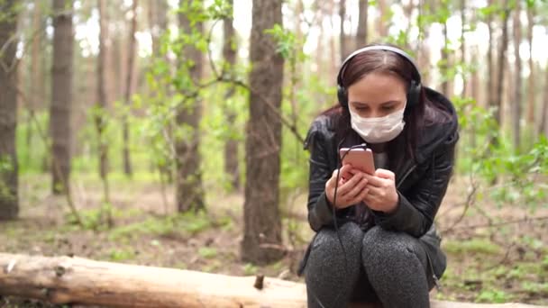 Primo piano di giovane donna in maschera medica ascoltare musica e sfogliare le notizie nel telefono cellulare, seduto sul log in foresta . — Video Stock