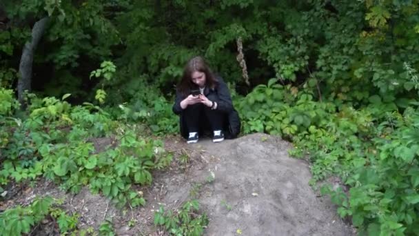 Woman sitting in the woods in windy weather and typing a message on a smartphone — Stock Video