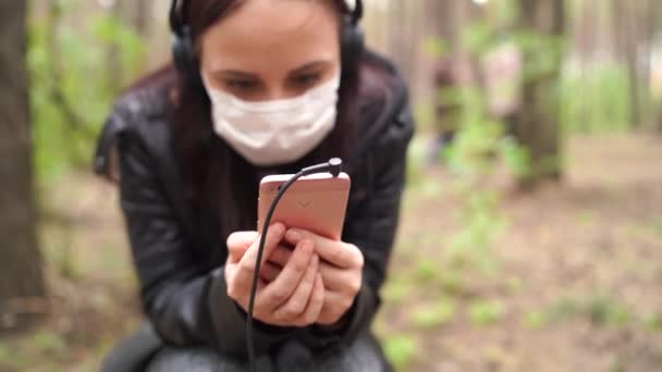 Närbild av ung kvinna i medicinsk mask lyssna på musik och bläddra igenom nyheter i mobiltelefon, sitter på log i skogen. — Stockvideo