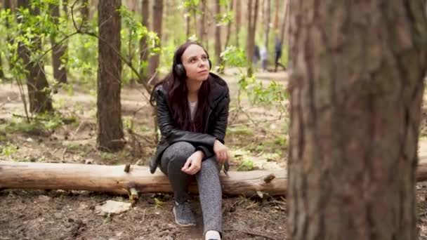 Glad ung kvinna lyssnar på musik, sitter på stocken i skogen. Vuxen kvinna njuter av sin vila på frisk luft. — Stockvideo