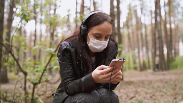 Primo piano di giovane donna in maschera medica ascoltare musica e sfogliare le notizie nel telefono cellulare, seduto sul log in foresta . — Video Stock