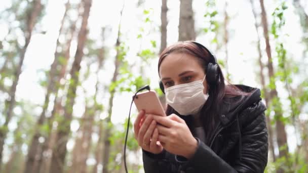 Närbild av ung kvinna i medicinsk mask lyssna på musik och bläddra igenom nyheter i mobiltelefon, sitter på log i skogen. — Stockvideo