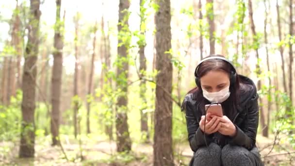 Zbliżenie młodej kobiety w masce medycznej słuchającej muzyki i przeglądającej wiadomości w telefonie komórkowym, siedzącej na logu w lesie. — Wideo stockowe