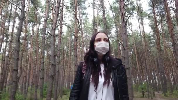 Yüzünde sağlık maskesi olan genç bir kadın temiz havada ormanda yürüyor. Yetişkin bir kadın kendini hastalıklardan korumak için yüzünü maskeyle örttü. Koronavirüs salgını tehdidi kavramı. — Stok video