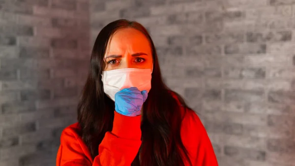 Молодая женщина в медицинской маске и перчатках кашляет в кулак, будучи изолированной дома. Взрослая женщина защищает себя от болезней. Концепция угрозы коронавирусной эпидемии . — стоковое фото