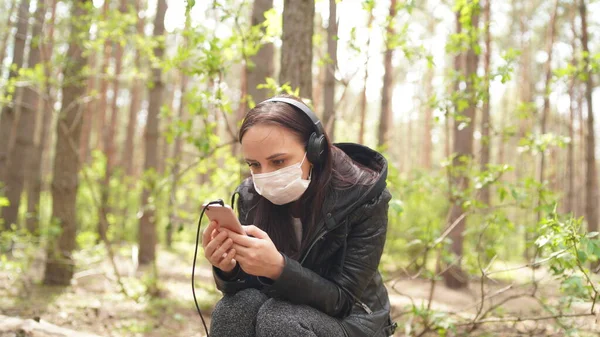 Крупный план молодой женщины в медицинской маске, слушающей музыку и листающей новости в мобильном телефоне, сидящей на логе в лесу . — стоковое фото