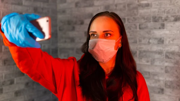 Молодая женщина в медицинской маске и перчатках делает селфи на смартфоне, стоя на сером кирпичном фоне. Портрет женщины, проводящей выходные дома и фотографирующей себя на мобильный телефон . — стоковое фото