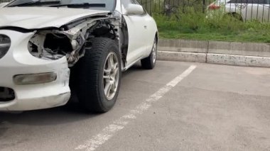 Kazadan sonra kaputu ve tamponu çökmüş beyaz bir araba. Asfaltlı park yerine park etmiş buruşuk metal şapkalı eski bir araba.