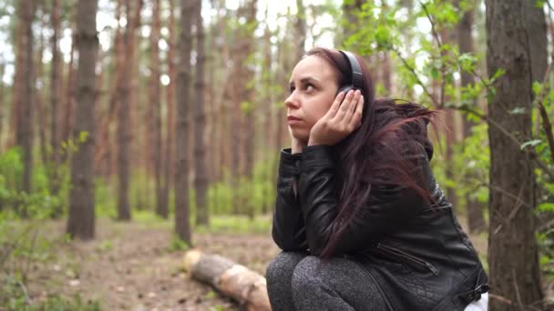 Happy young woman listening to music, sitting on log in forest. Adult female enjoying her rest on fresh air. — Stock Video