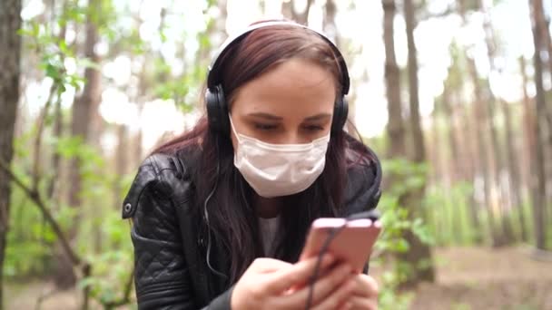 Närbild av ung kvinna i medicinsk mask lyssna på musik och bläddra igenom nyheter i mobiltelefon, sitter på log i skogen. — Stockvideo