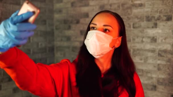 Jeune femme en masque médical et gants prenant selfie sur smartphone, debout sur fond de brique grise. Portrait d'une femme passant un week-end à la maison et se photographiant sur un téléphone portable . — Video