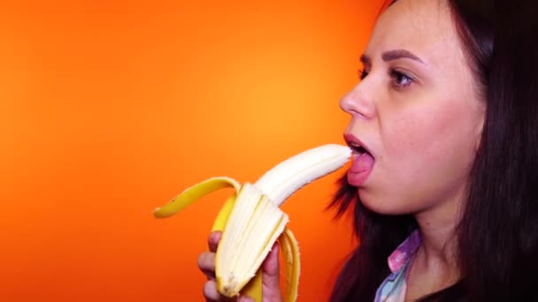 Close-up van mooie jonge vrouw die banaan eet op oranje achtergrond. volwassen brunette bijt het fruit en kauwt, weg te kijken. — Stockvideo