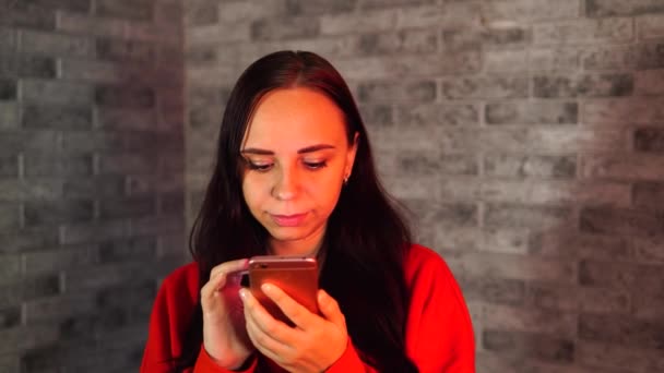 Ładna młoda kobieta w czerwonej bluzie z kapturem odwraca wiadomości o wirusie w telefonie komórkowym, stojąca na szarym tle z cegły. — Wideo stockowe