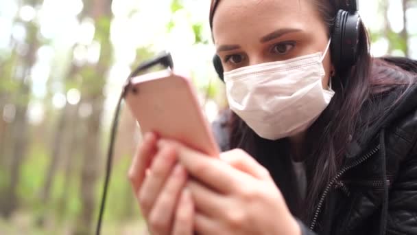 Nahaufnahme einer jungen Frau in medizinischer Maske, die Musik hört und im Handy Nachrichten durchblättert, auf einem Baumstamm im Wald sitzt. — Stockvideo