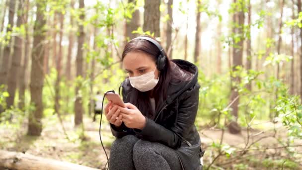 Primo piano di giovane donna in maschera medica ascoltare musica e sfogliare le notizie nel telefono cellulare, seduto sul log in foresta . — Video Stock