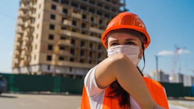 Tulumlu ve tıbbi maskeli kadın inşaat halindeki evin arka planında dirsek öksürüyor. Koronavirüs salgını tehdidi kavramı.