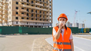 Tulumlu ve tıbbi maskeli kadın inşaat halindeki evin arka planında öksürüp duruyor. Koronavirüs salgını tehdidi kavramı.