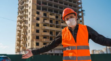 İnşaat halindeki evin arka planında tıbbi maskeli ve tulumlu erkek inşaat işçisinin portresi..