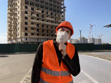 İnşaat halindeki evin arka planında tıbbi maskeli ve tulumlu erkek inşaat işçisinin portresi..