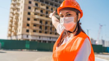 Tulumlu ve tıbbi maskeli kadın inşaat halindeki evi işaret ediyor..