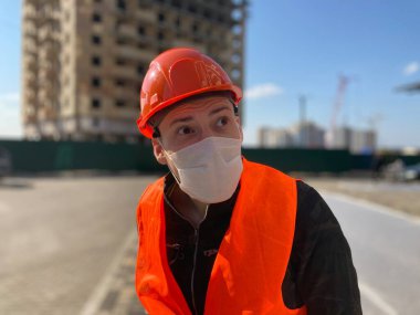 İnşaat halindeki evin arka planında tıbbi maskeli ve tulumlu erkek inşaat işçisinin portresi..