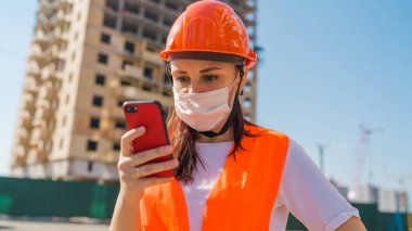 Tulumlu ve tıbbi maskeli kadın inşaat halindeki evin arka planındaki cep telefonunda Coronavirus 'la ilgili haberleri inceliyor. Enfeksiyon tehdidi kavramı.