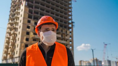 İnşaat halindeki evin arka planında tıbbi maskeli ve tulumlu erkek inşaat işçisinin portresi..