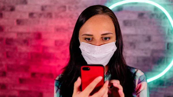 Joven hermosa mujer en la máscara médica se para con el teléfono móvil en las manos en la pared de ladrillo de fondo con lámparas brillantes. Retrato de encantadora mujer hojeando varias noticias en el teléfono móvil contra — Foto de Stock