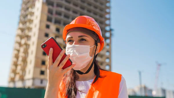 Female construction worker in overalls and medical mask flipping through news about coronavirus in mobile phone on background of house under construction. Concept of threat of infection.