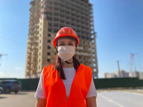 Portrait of female construction worker in medical mask and overalls on background of house under construction.