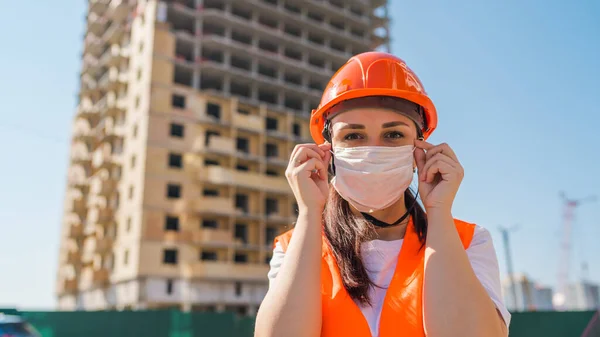 Female construction worker in overalls putting on medical mask on face on background of house under construction.