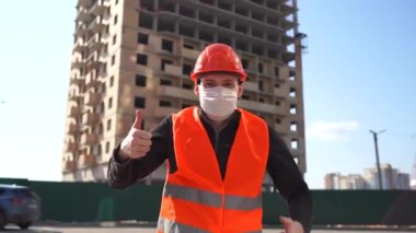Tulumlu ve tıbbi maskeli bir erkek inşaat halindeki evin arka planında onay gösteriyor. Sert şapkalı ve turuncu yelekli genç adam onay jesti yapıyor..
