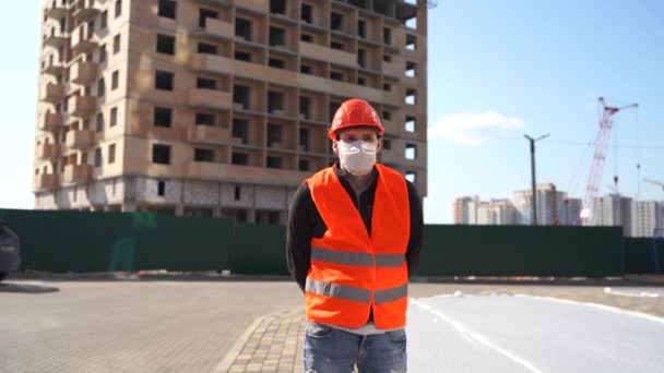 Retrato del trabajador de la construcción masculino en máscara médica y overoles sobre fondo de casa en construcción . — Vídeos de Stock