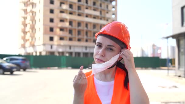 Žena stavební dělnice v kombinézách nasazení lékařské masky na tvář na pozadí domu ve výstavbě. — Stock video