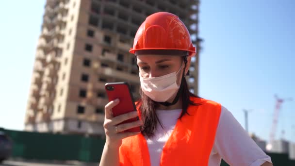 Kobieta pracownik budowlany w kombinezon i medyczny maska, przedzieram się przez nowina o koronawirus w komórkowy telefon na tle budowlany dom. Pojęcie zagrożenia zakażeniem. — Wideo stockowe