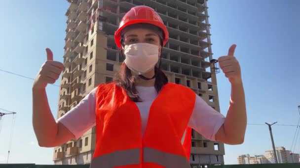 Женщина-строитель в комбинезоне и в медицинской маске показывает большие пальцы на фоне строящегося дома. Молодая женщина в каске и оранжевом жилете демонстрирует жест одобрения . — стоковое видео