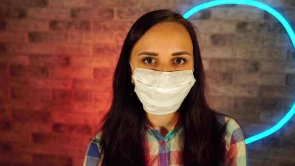 Молодая женщина в медицинской маске позирует с мобильным телефоном на фоне обветшалой кирпичной стены с светящимися лампами. Портрет очаровательной женщины у подсвеченной стены . — стоковое видео