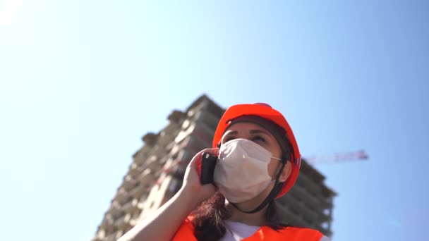 Женщина-строитель в комбинезоне и медицинской маске говорит о коронавирусе на мобильном телефоне на фоне строящегося дома. Концепция угрозы заражения . — стоковое видео