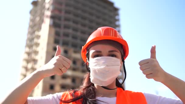 Female construction worker in overalls and in medical mask showing thumbs up on background of house under construction. Young woman in hard hat and orange vest showing gesture of approval. — Stock Video