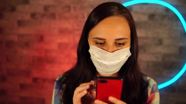 Νεαρή όμορφη γυναίκα στην ιατρική μάσκα στέκεται με κινητό τηλέφωνο στα χέρια σε τοίχο από τούβλα φόντο με λαμπερό λαμπτήρες. Πορτρέτο του γοητευτικό θηλυκό flipping μέσα από διάφορες ειδήσεις στο κινητό τηλέφωνο κατά — Αρχείο Βίντεο